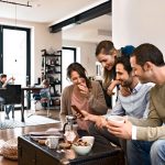 4 fröhliche junge Menschen sitzen in einem offenen Raum an einem Tisch und betrachten etwas auf einem Smartphone.