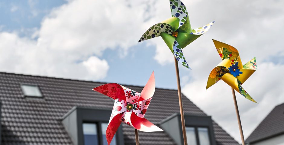 Drei bunte Kinderwindmühlen drehen sich im Wind. Dahinter ist ein Haus zu sehen.