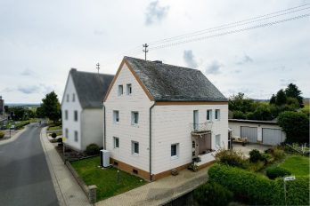 : In einer ländlichen Umgebung stehen zwei zweistöckige Einfamilienhäuser an einer Hauptstraße.