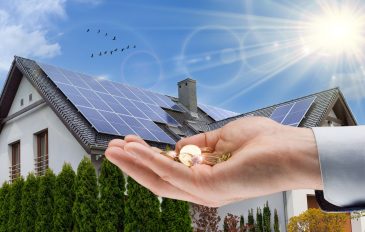 Eine ausgestreckte Hand, gefüllt mit Euromünzen, vor einem Haus mit installierten Photovoltaikmodulen. Im Hintergrund scheint die Sonne, der Himmel ist blau