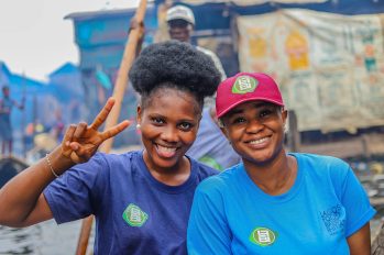 Zwei Frauen aus Nigeria lächeln in die Kamera, im Hintergrund ein Slum