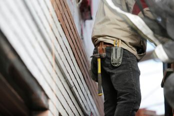 Ein angeschnittener Bauarbeiter steht mit einem Hammer in seinem Holster vor einer Hauswand.